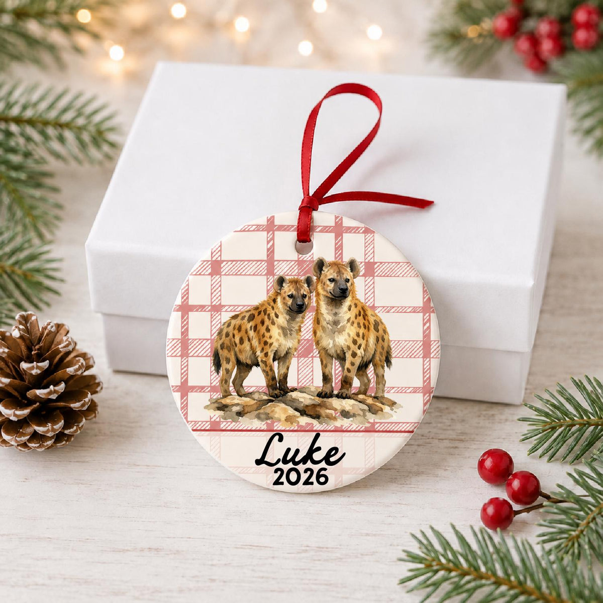 Personalized spotted hyena ceramic ornament with name and year on red plaid in front of a white gift box with pinecones and red berries