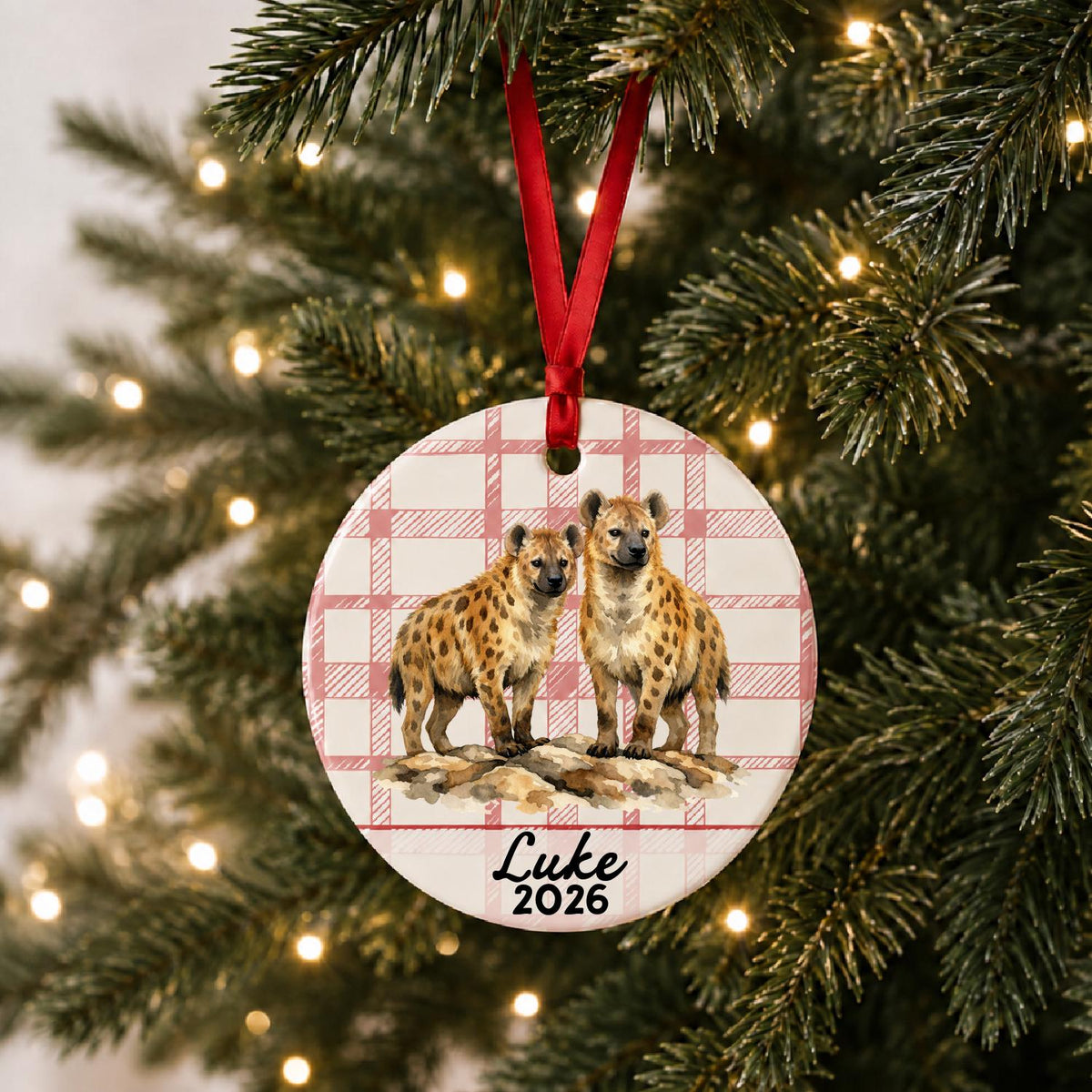 Personalized spotted hyena ceramic ornament with name and year on red plaid hanging on a lit Christmas tree