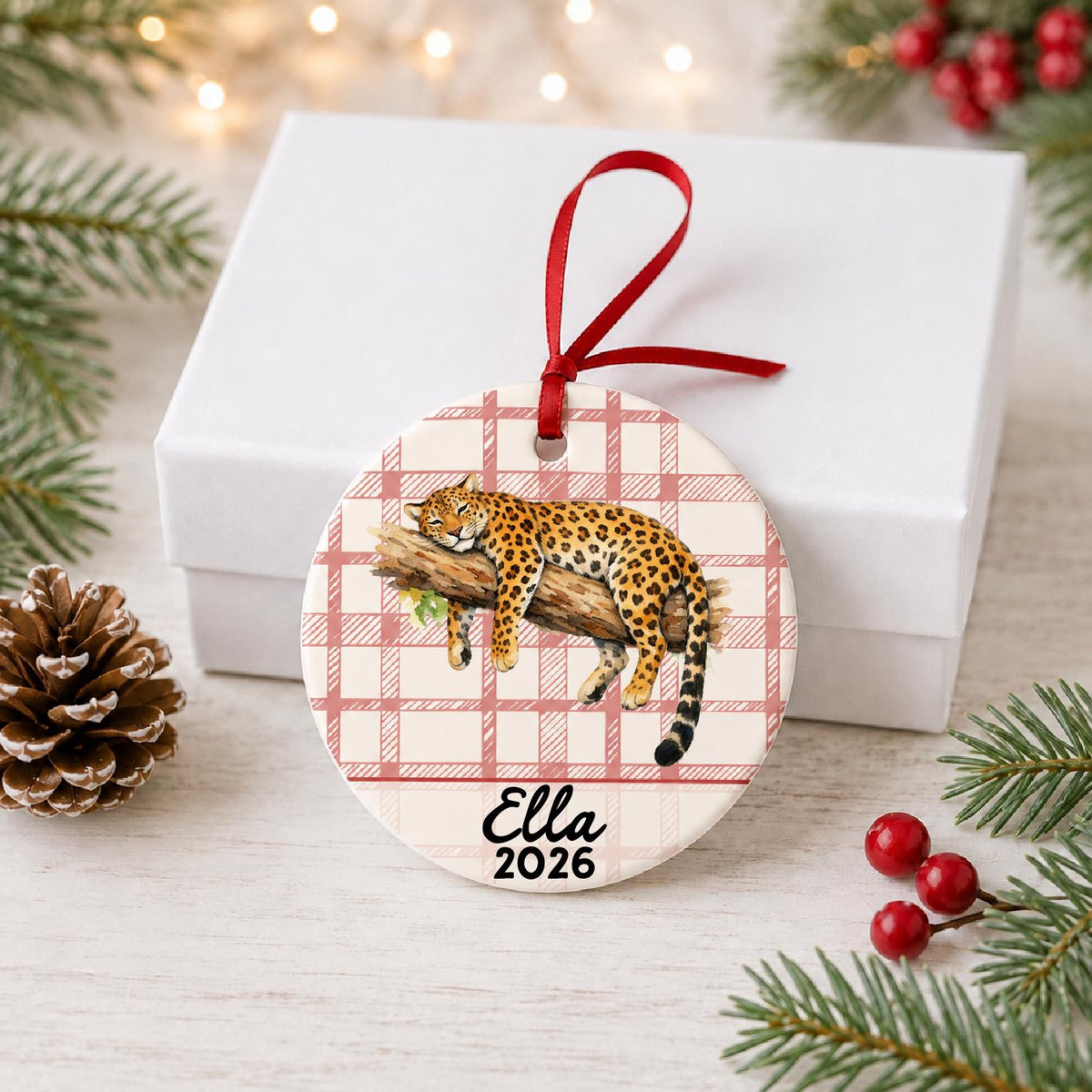 Personalized leopard ceramic ornament with name and year on red plaid in front of a white gift box with pinecones and red berries