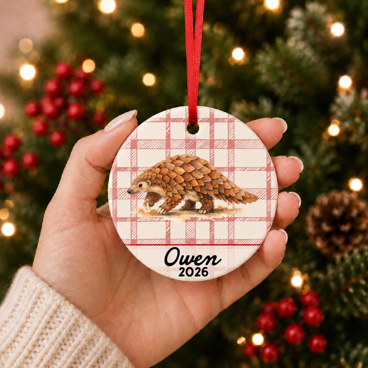 Hand holding the personalized pangolin ceramic ornament with name and year on red plaid in front of a Christmas tree
