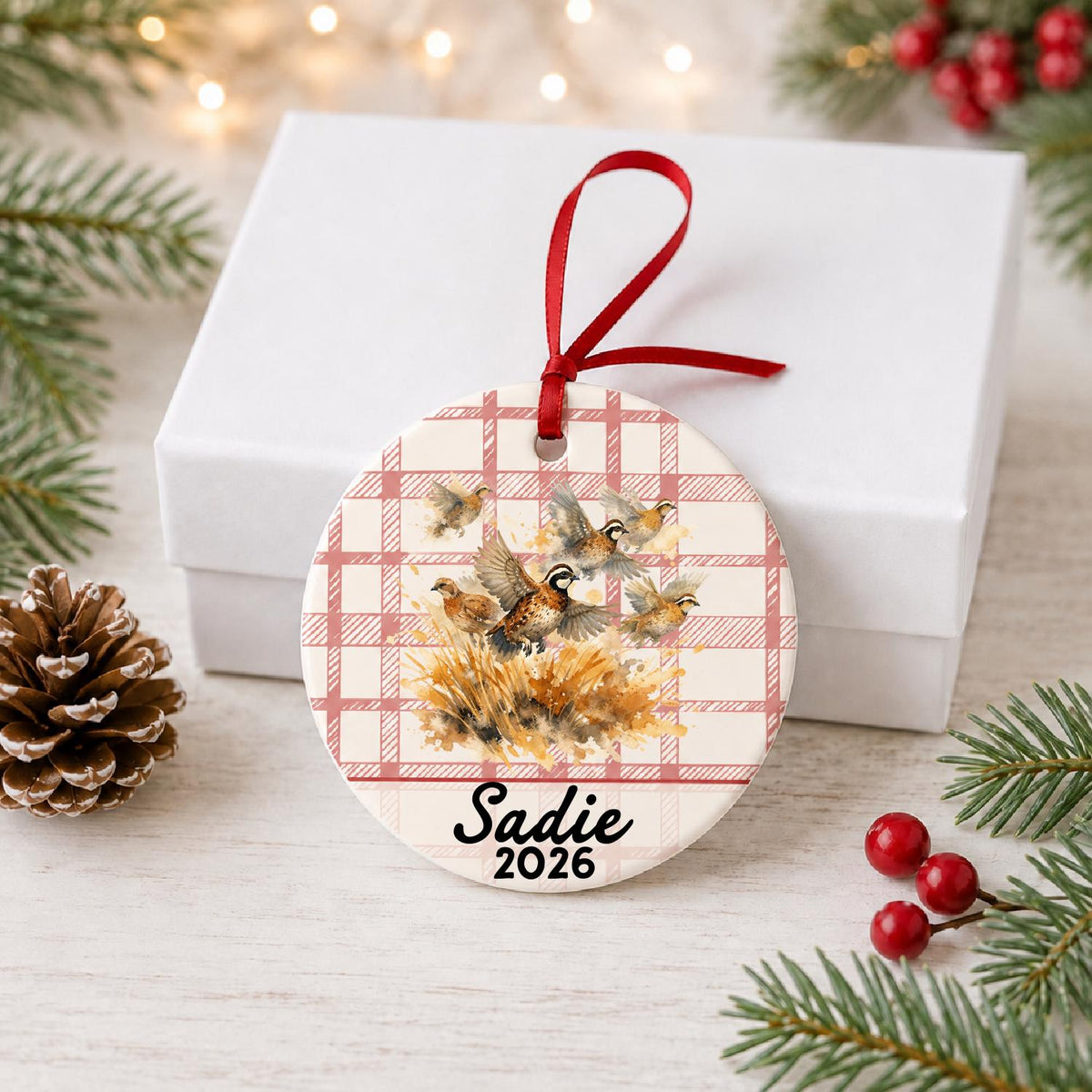 Personalized bobwhite quail ceramic ornament with name and year on red plaid in front of a white gift box with pinecones and red berries