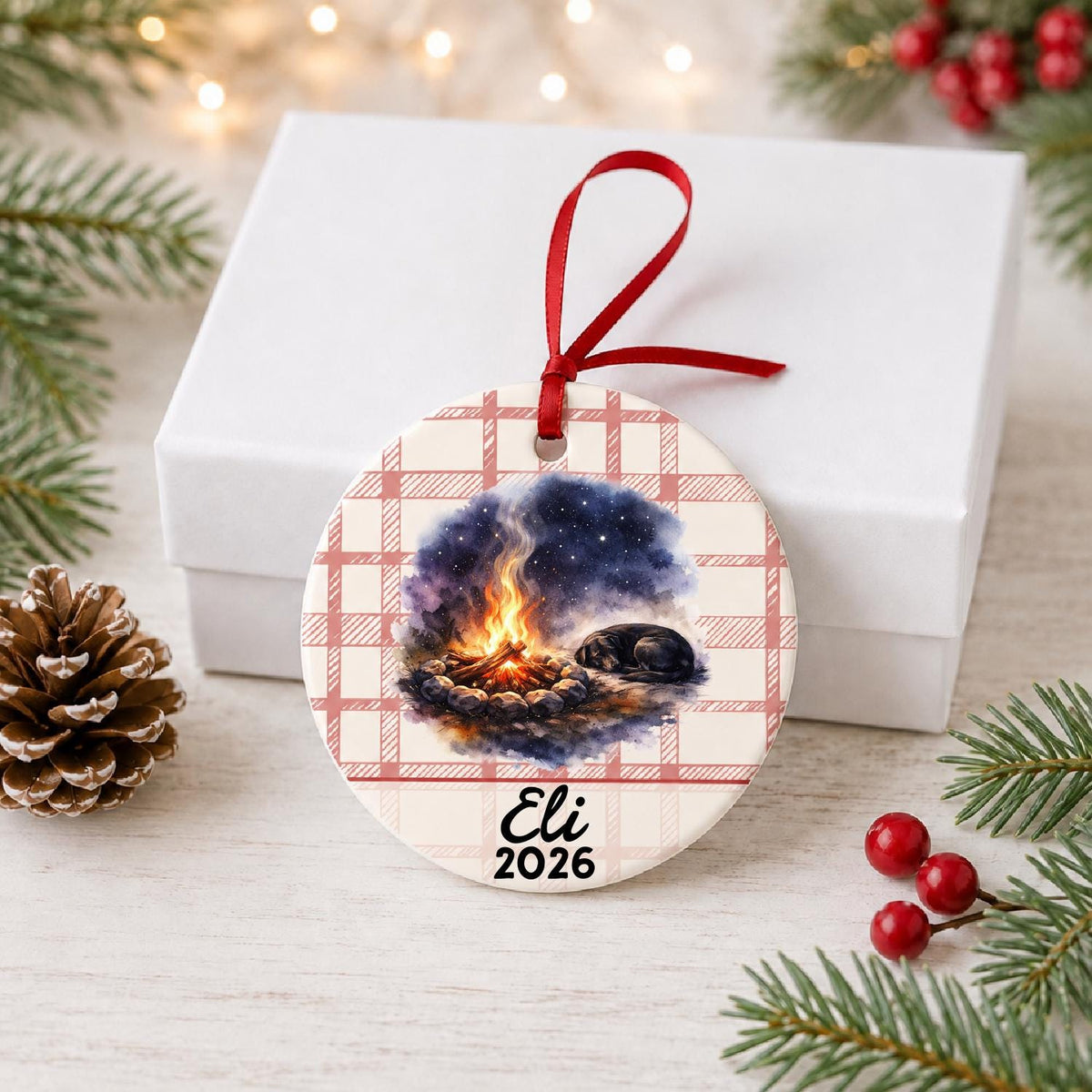 Personalized campfire and dog ceramic ornament with name and year on red plaid in front of a white gift box with pinecones and red berries