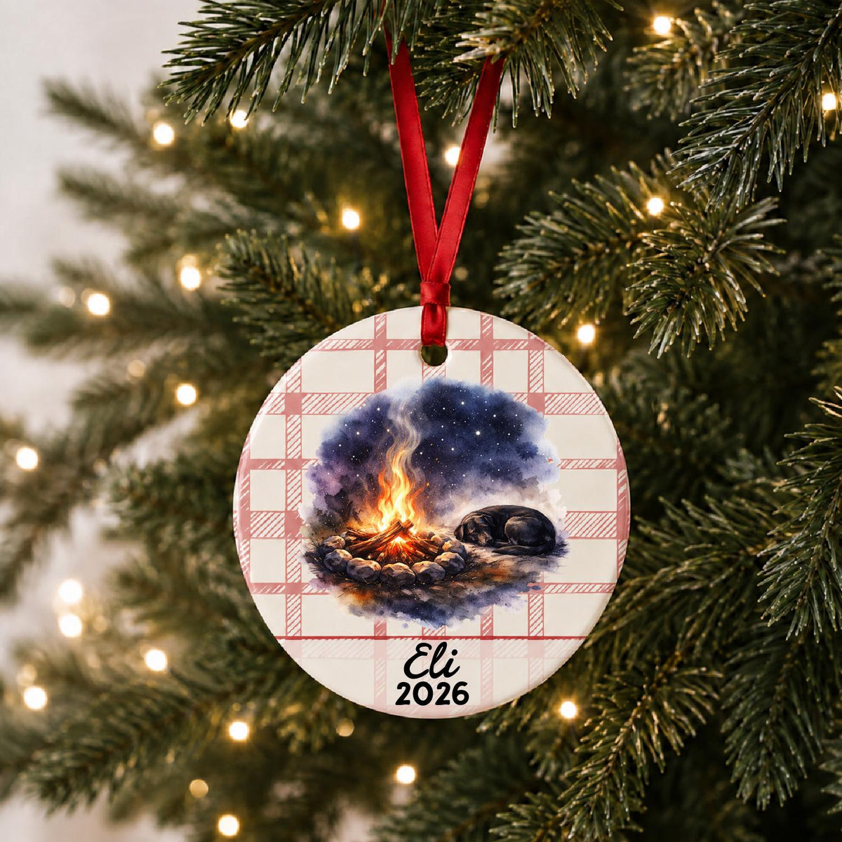 Personalized campfire and dog ceramic ornament with name and year on red plaid hanging on a lit Christmas tree