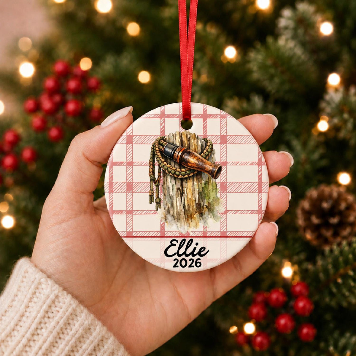Hand holding the personalized duck call ceramic ornament with name and year on red plaid in front of a Christmas tree