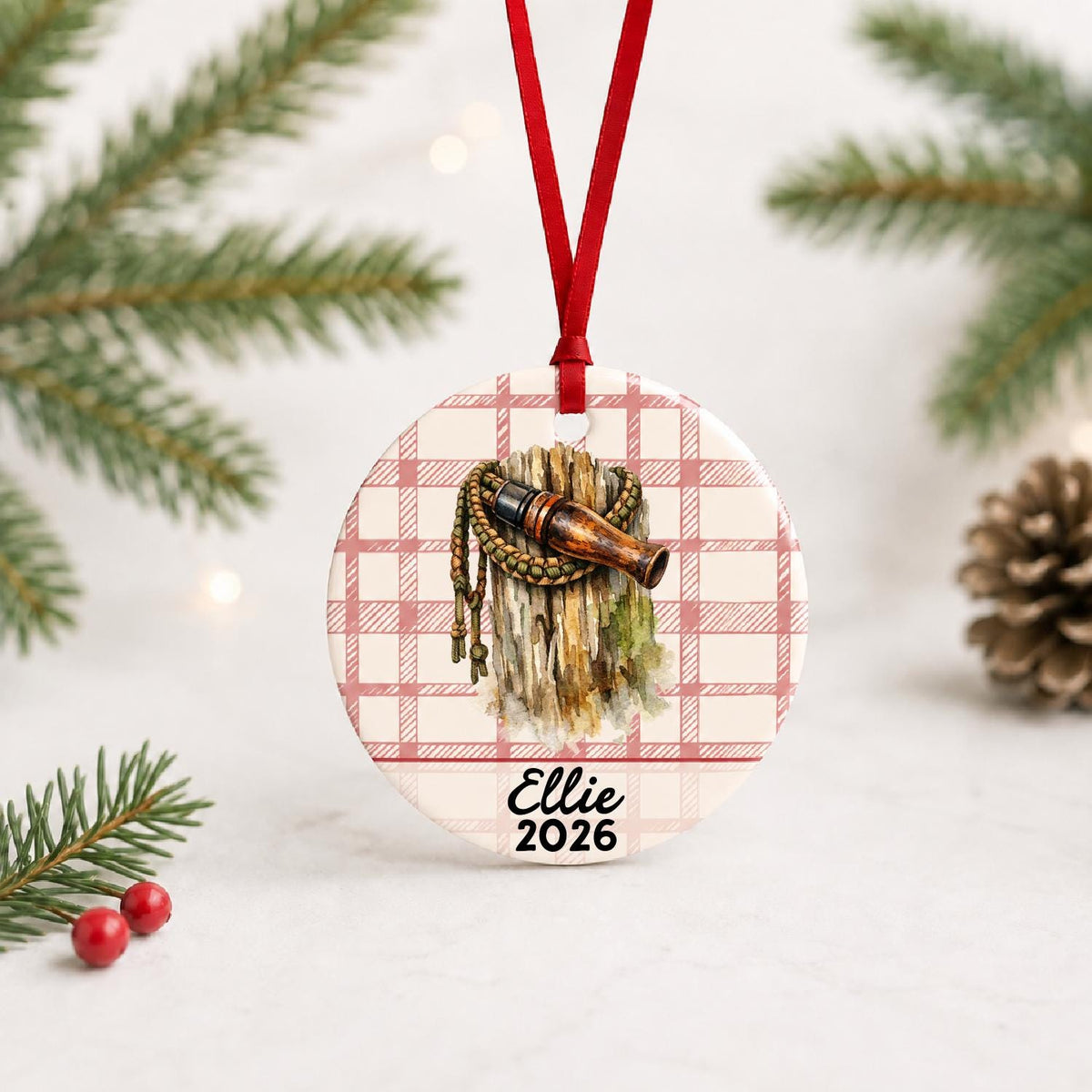 Personalized duck call ceramic ornament with name and year on red plaid with red ribbon on a white surface with pine branches and pinecones