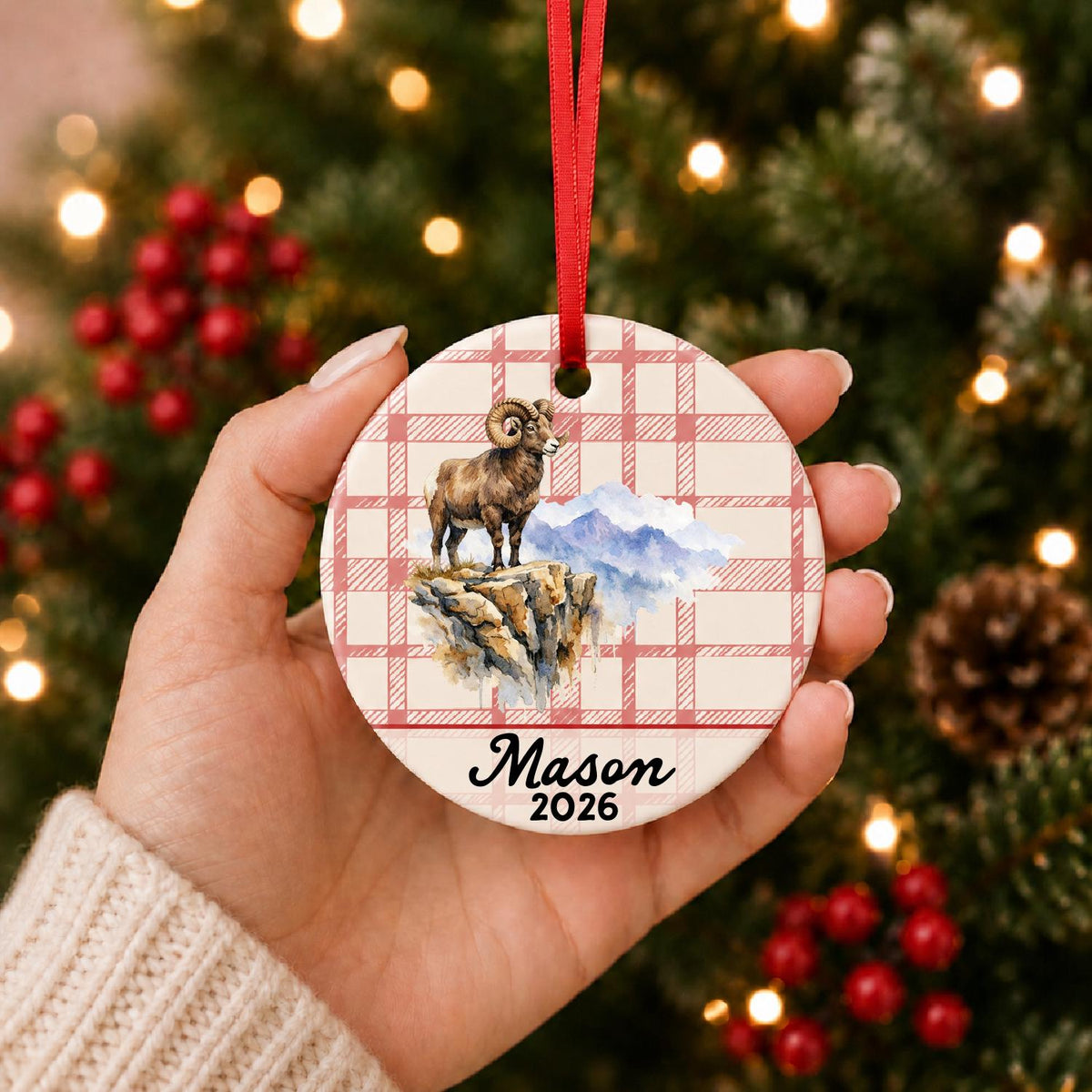 Hand holding the personalized bighorn sheep ceramic ornament with name and year on red plaid in front of a Christmas tree
