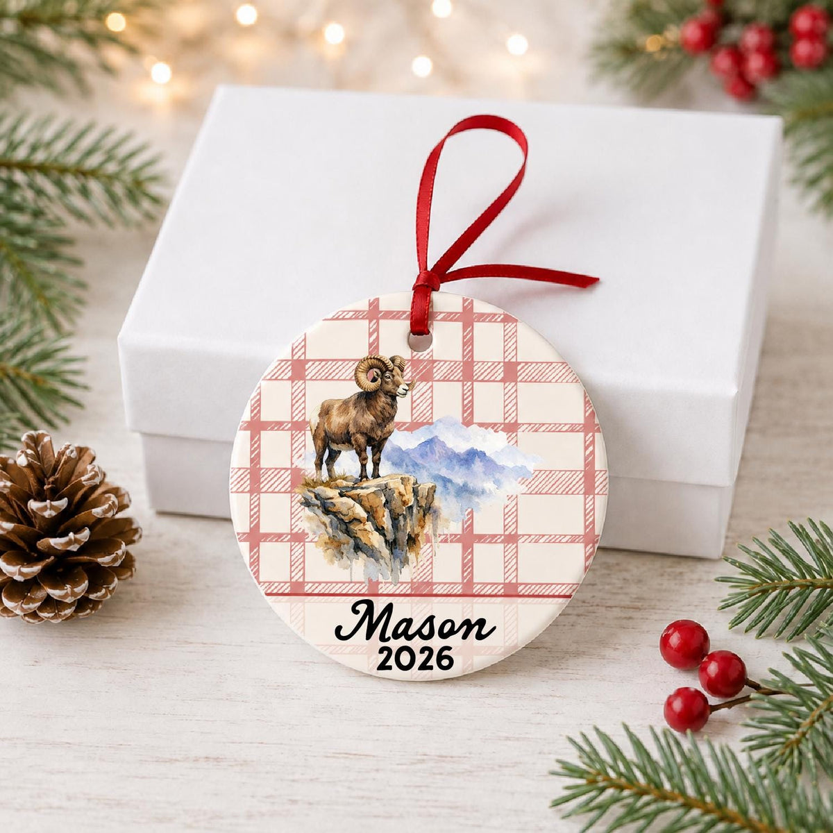 Personalized bighorn sheep ceramic ornament with name and year on red plaid in front of a white gift box with pinecones and red berries