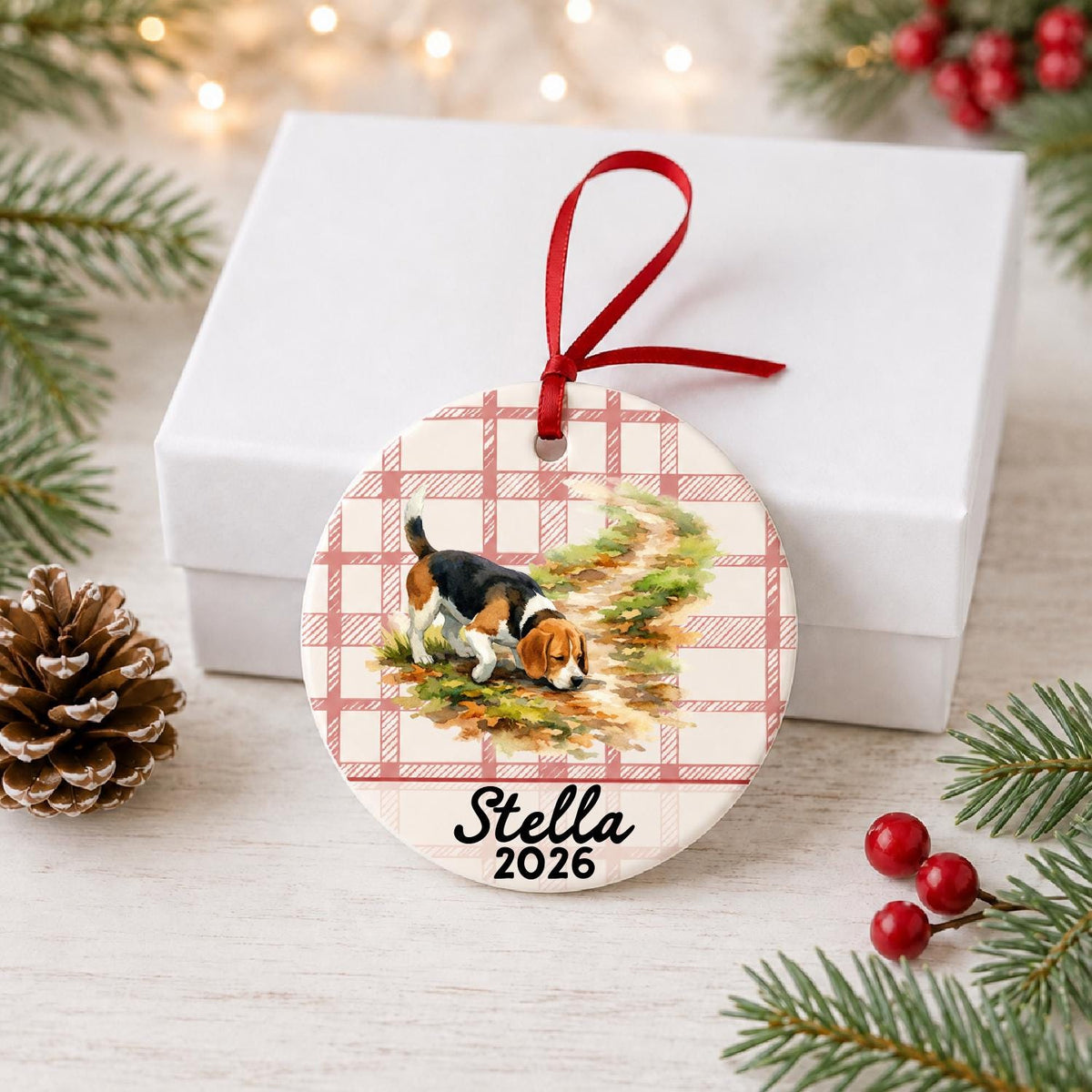 Personalized beagle ceramic ornament with name and year on red plaid in front of a white gift box with pinecones and red berries