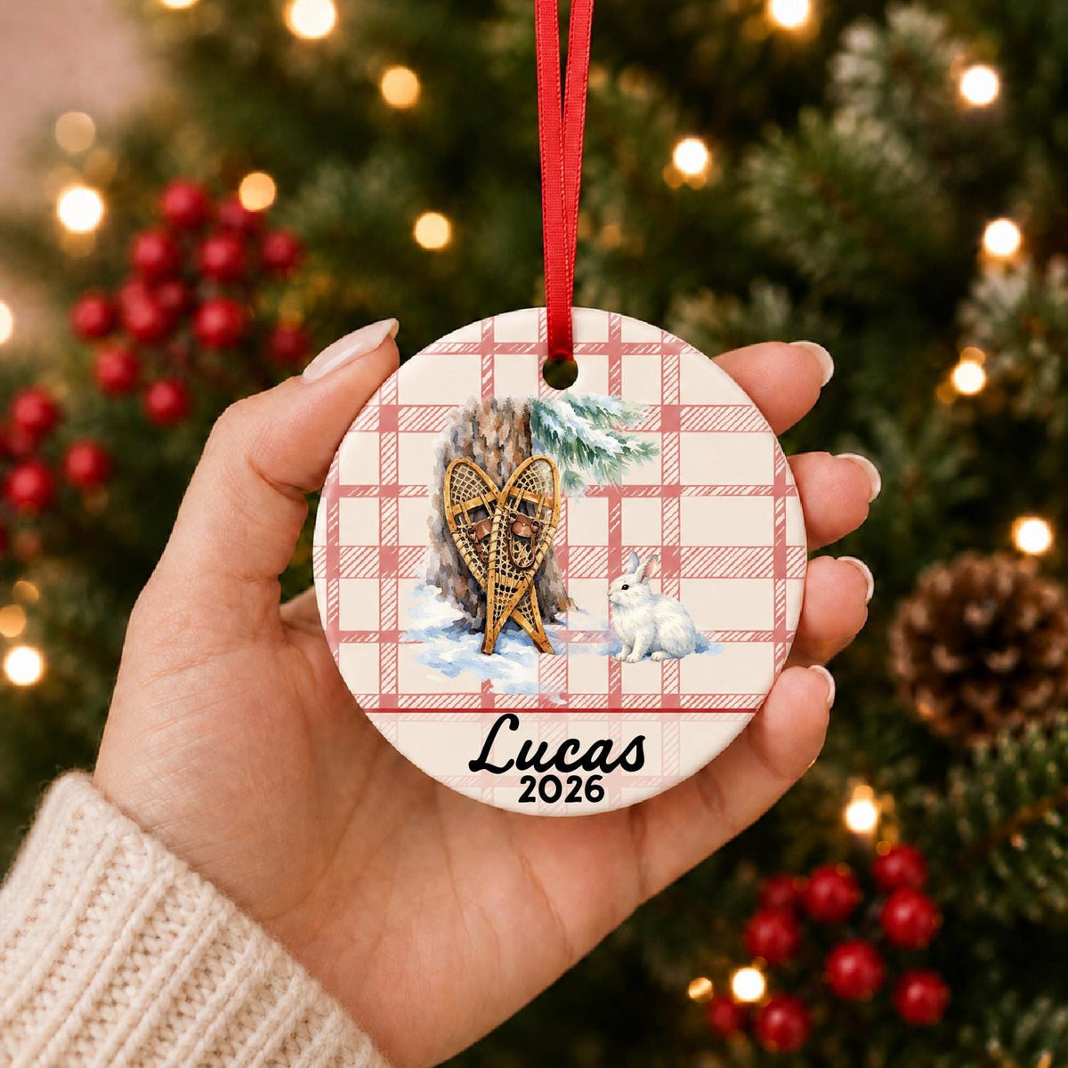 Hand holding the personalized snowshoes and snowshoe hare ceramic ornament with name and year on red plaid in front of a Christmas tree