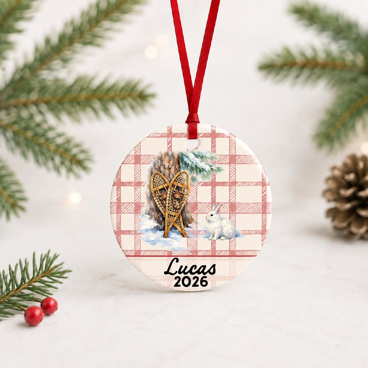Personalized snowshoes and snowshoe hare ceramic ornament with name and year on red plaid with red ribbon on a white surface with pine branches and pinecones