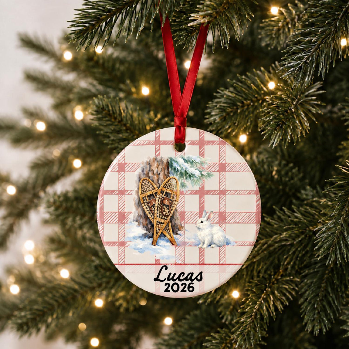 Personalized snowshoes and snowshoe hare ceramic ornament with name and year on red plaid hanging on a lit Christmas tree