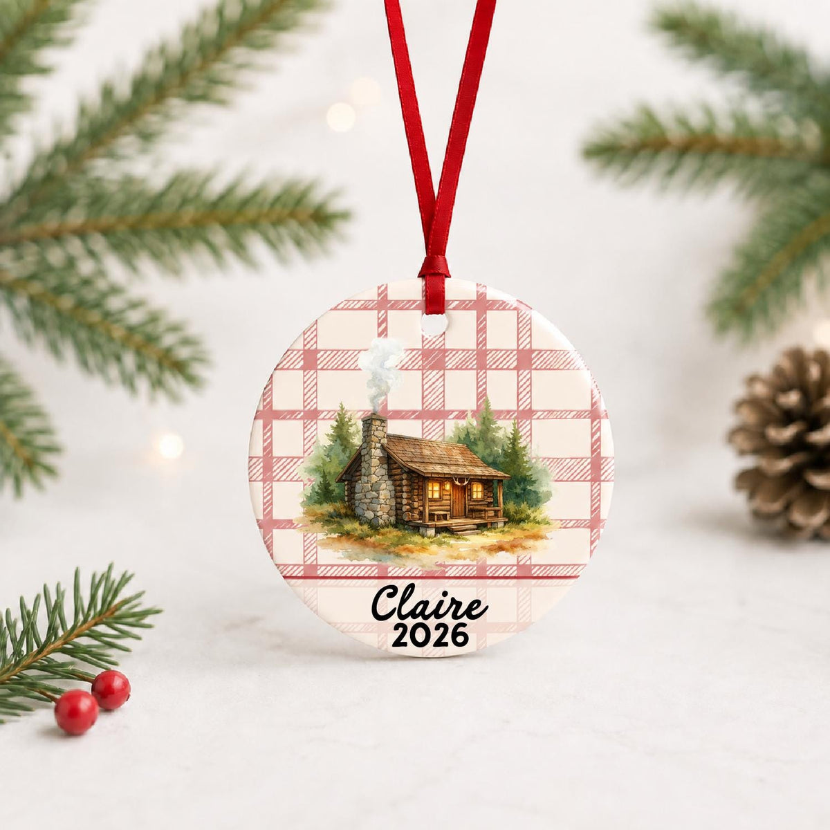 Personalized log cabin ceramic ornament with name and year on red plaid with red ribbon on a white surface with pine branches and pinecones