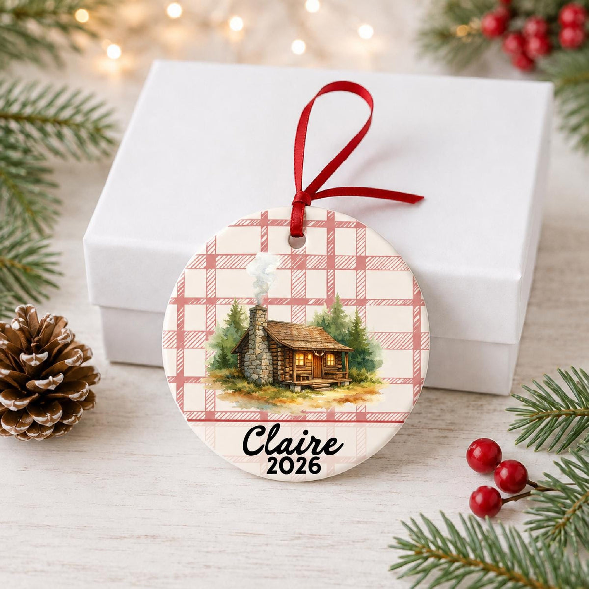 Personalized log cabin ceramic ornament with name and year on red plaid in front of a white gift box with pinecones and red berries