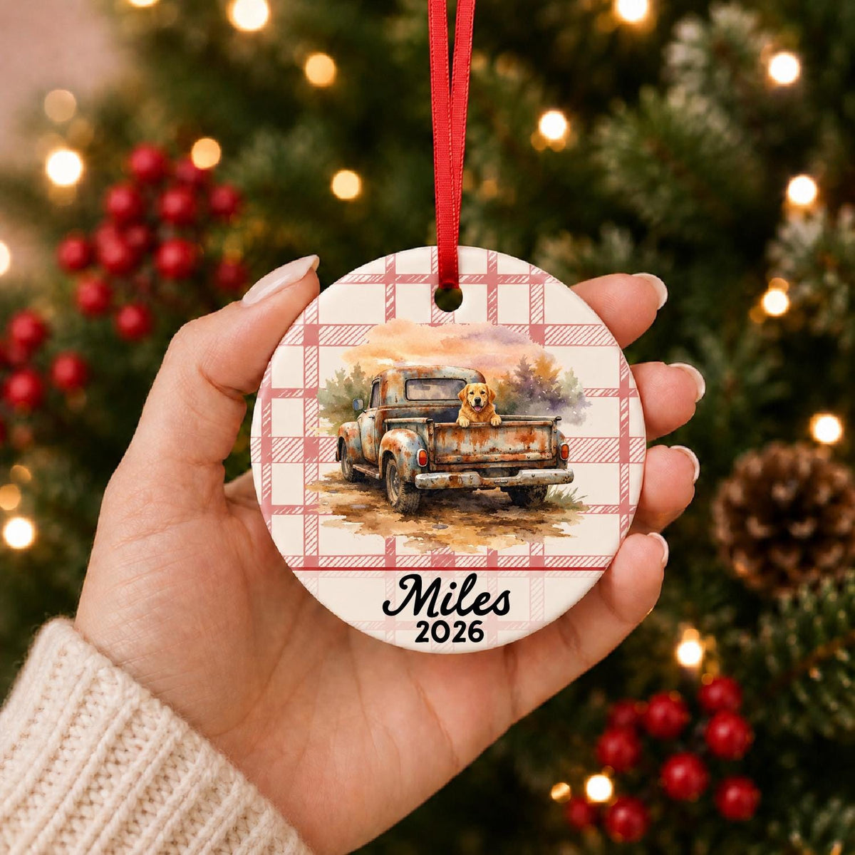 Hand holding the personalized vintage truck and dog ceramic ornament with name and year on red plaid in front of a Christmas tree