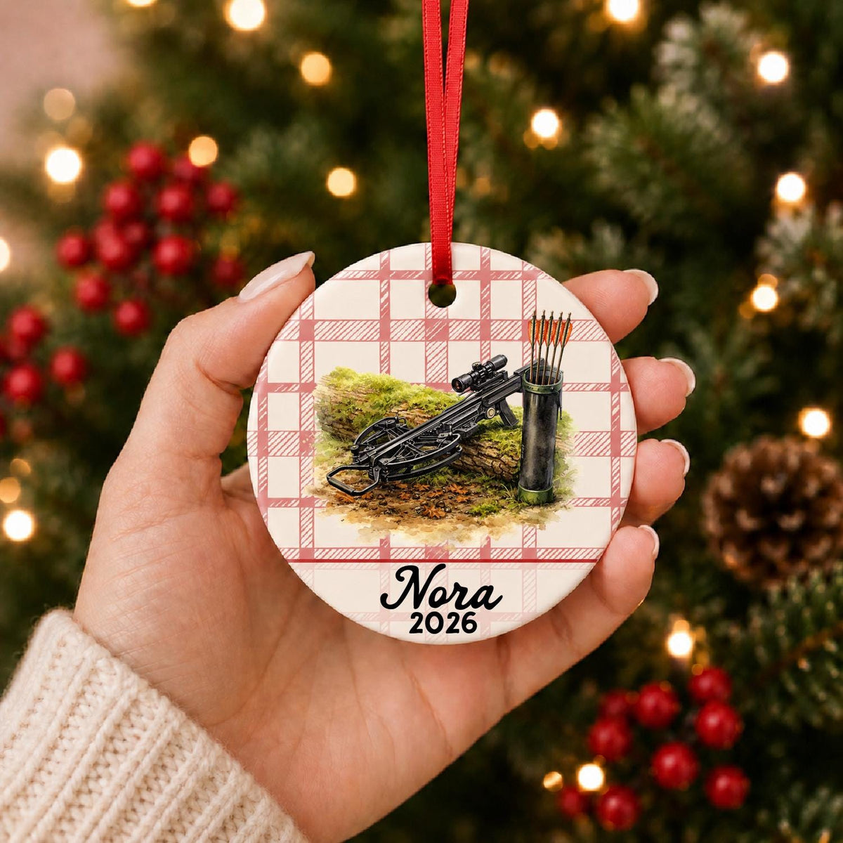 Hand holding the personalized crossbow ceramic ornament with name and year on red plaid in front of a Christmas tree