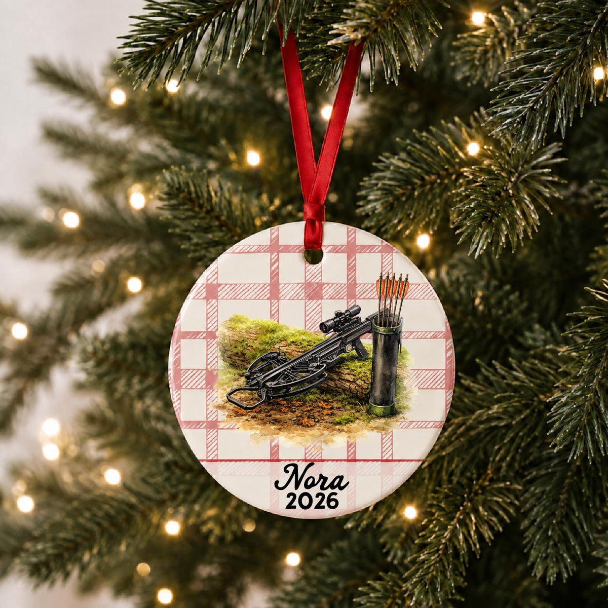 Personalized crossbow ceramic ornament with name and year on red plaid hanging on a lit Christmas tree