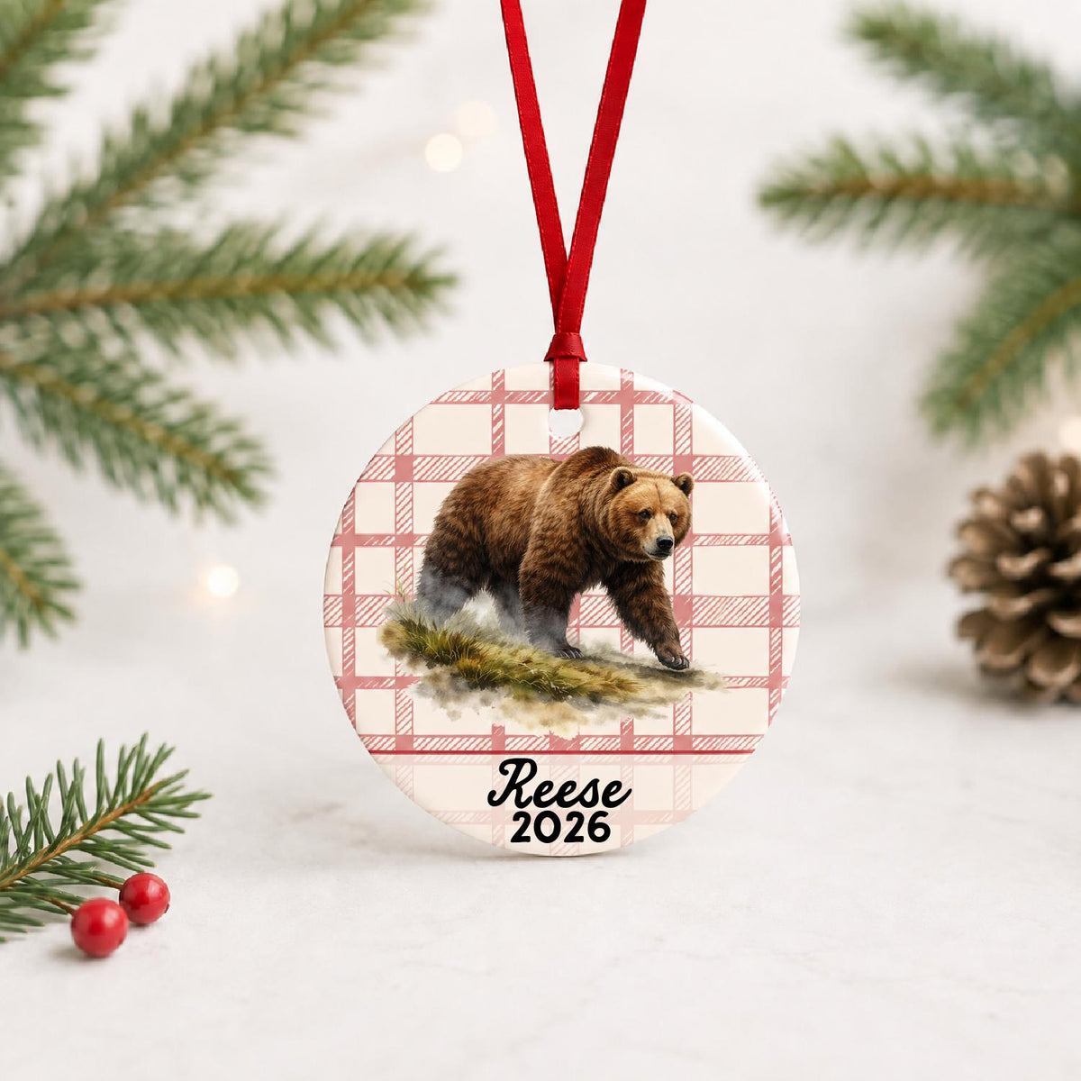 Personalized brown bear ceramic ornament with name and year on red plaid with red ribbon on a white surface with pine branches and pinecones