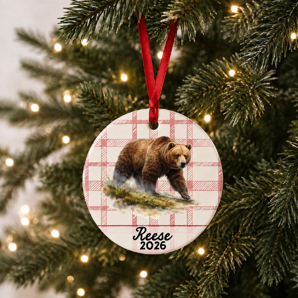 Personalized brown bear ceramic ornament with name and year on red plaid hanging on a lit Christmas tree