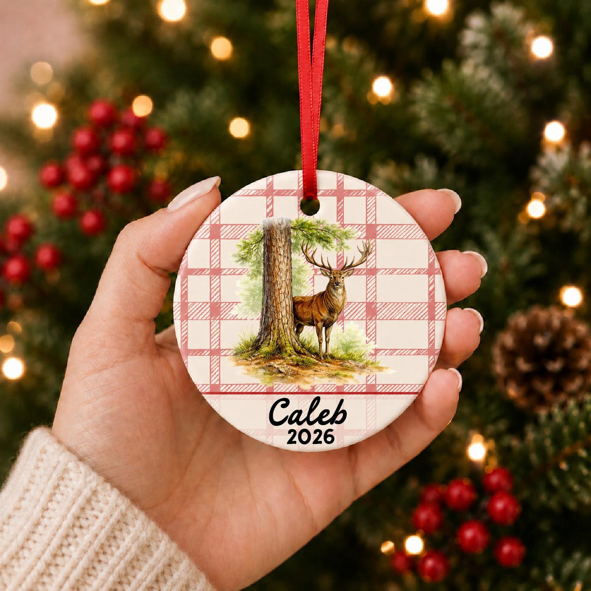 Hand holding the personalized red deer stag ceramic ornament with name and year on red plaid in front of a Christmas tree