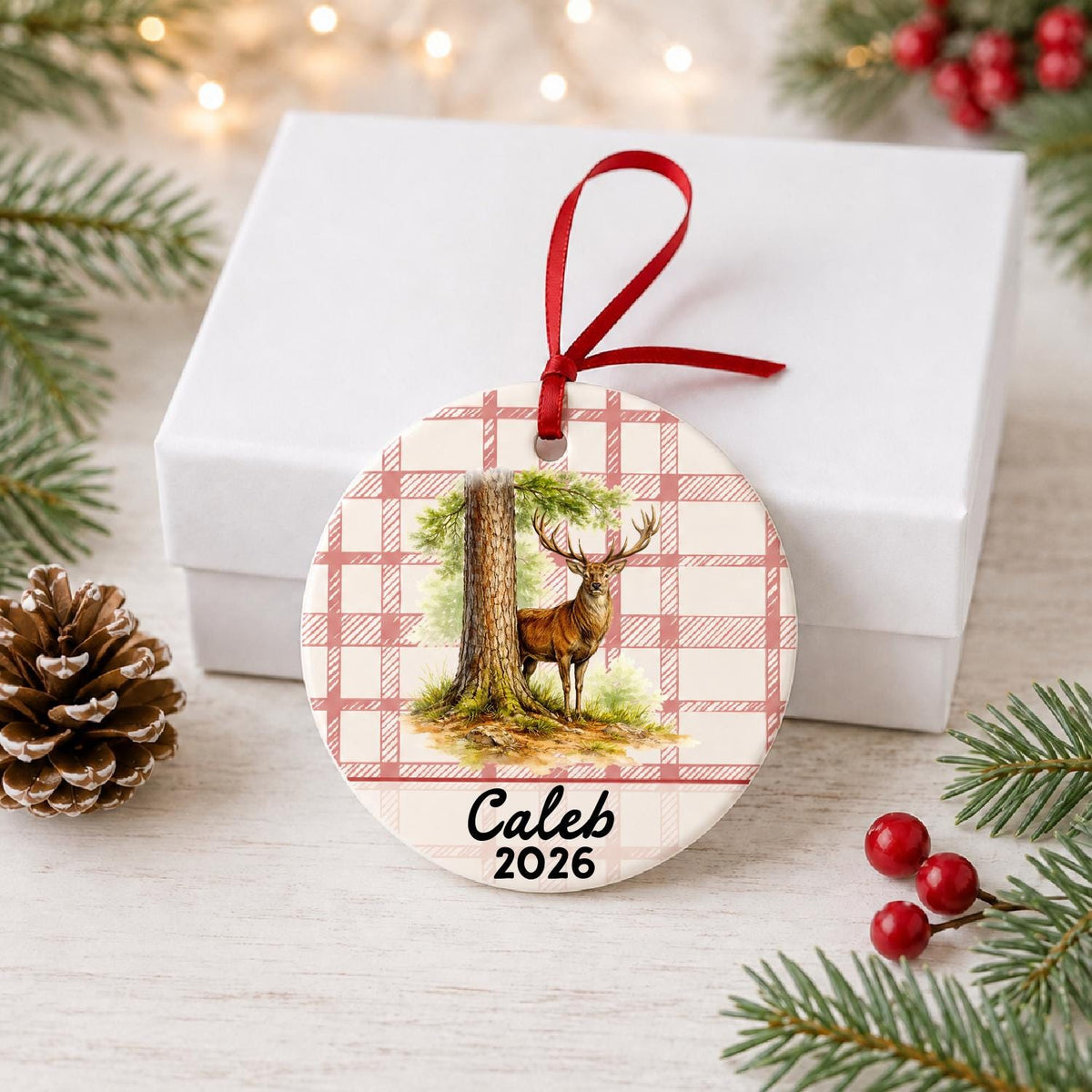 Personalized red deer stag ceramic ornament with name and year on red plaid in front of a white gift box with pinecones and red berries