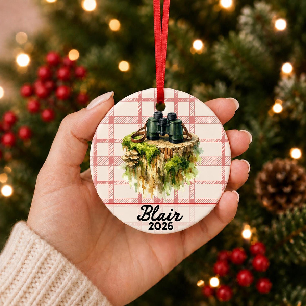 Hand holding the personalized binoculars ceramic ornament with name and year on red plaid in front of a Christmas tree
