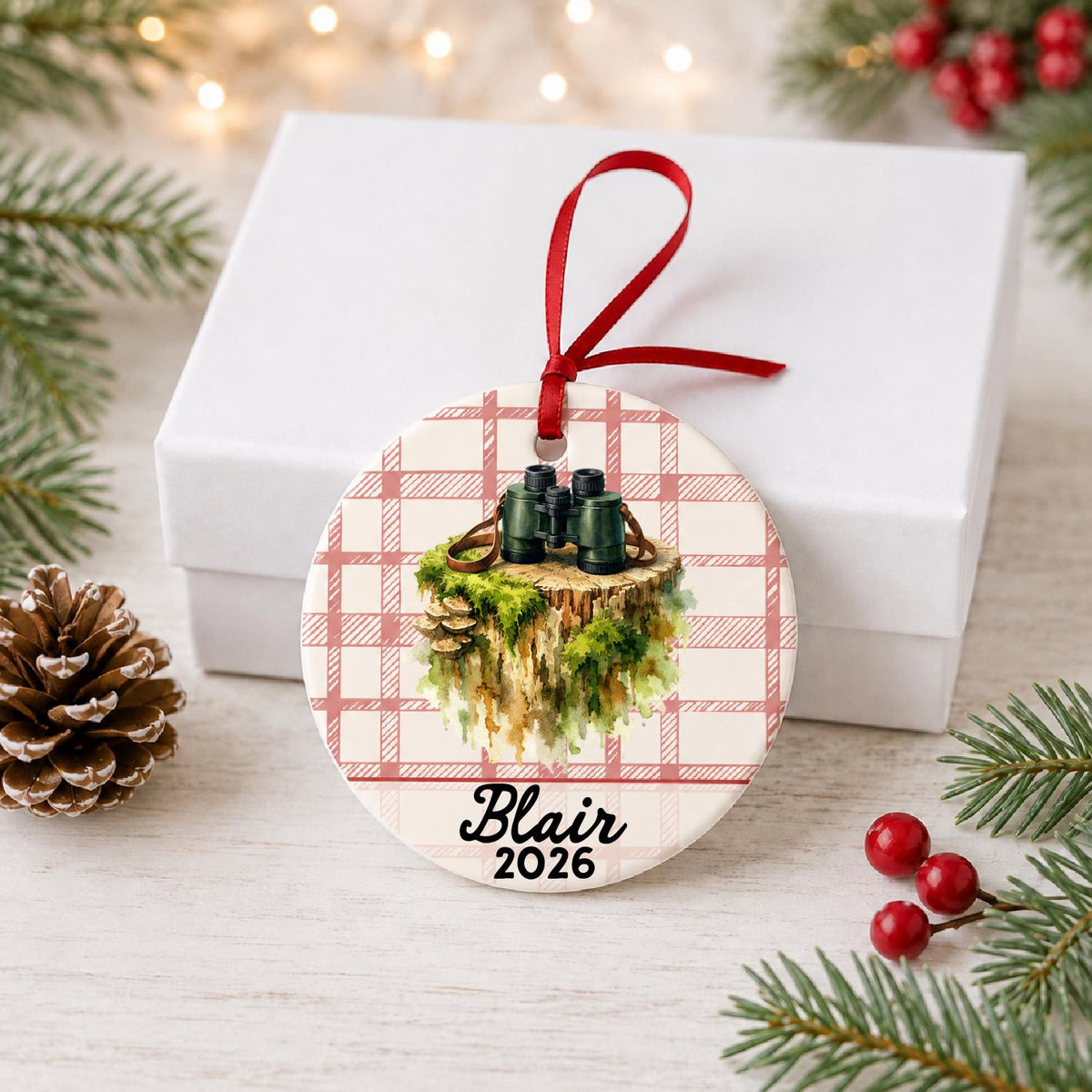 Personalized binoculars ceramic ornament with name and year on red plaid in front of a white gift box with pinecones and red berries