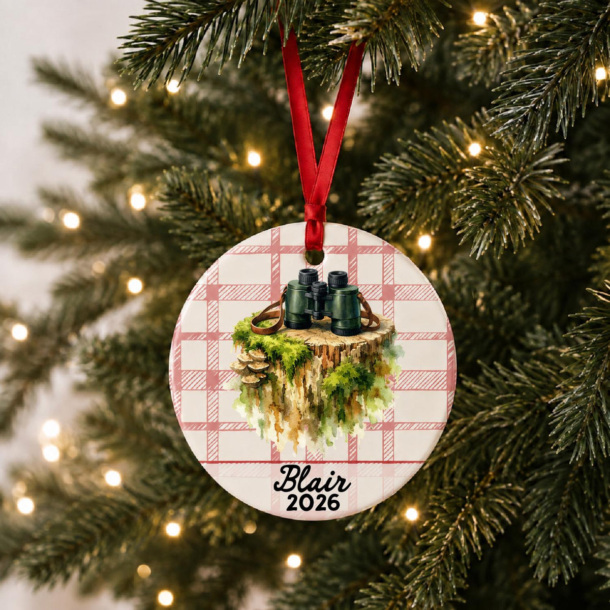 Personalized binoculars ceramic ornament with name and year on red plaid hanging on a lit Christmas tree