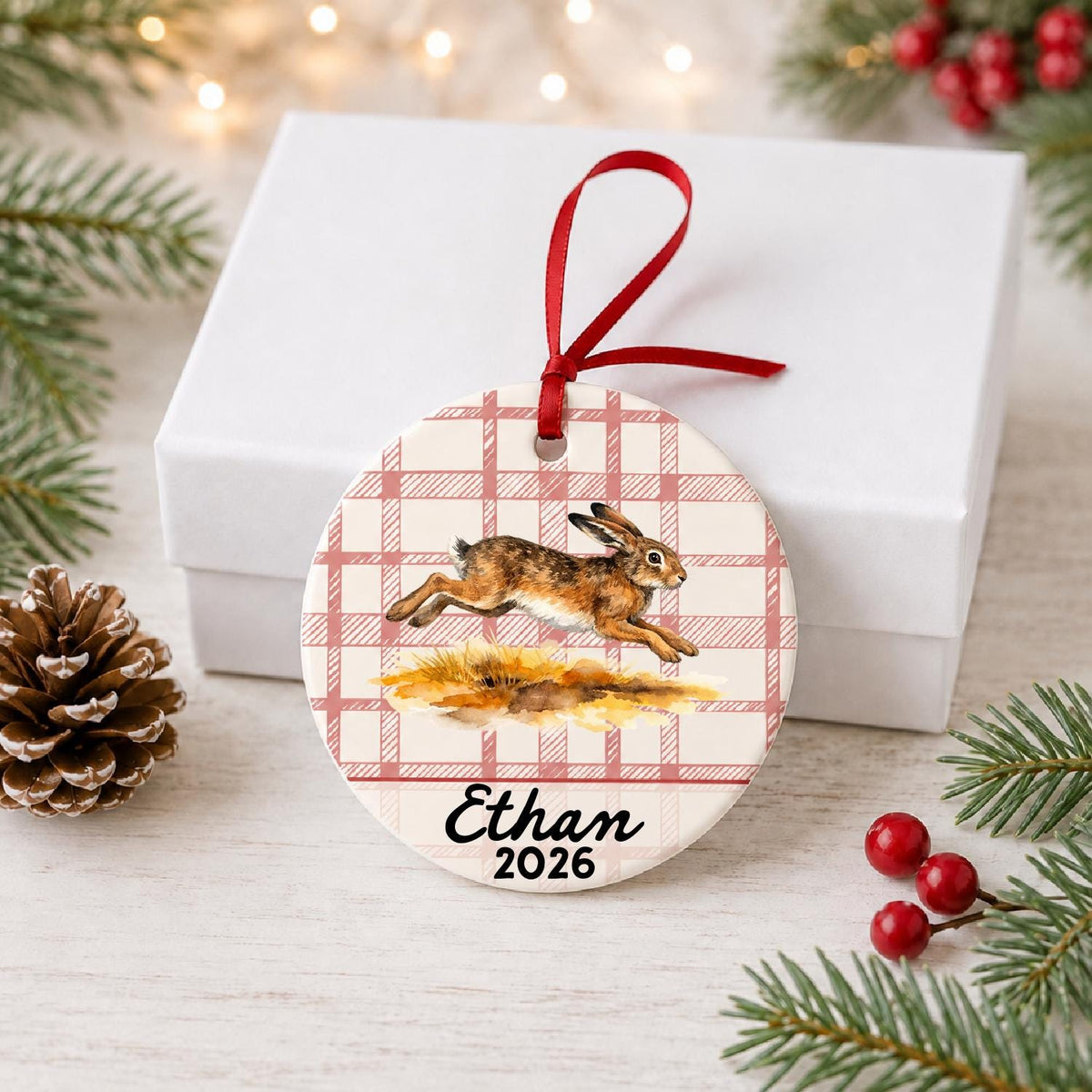 Personalized brown hare ceramic ornament with name and year on red plaid in front of a white gift box with pinecones and red berries