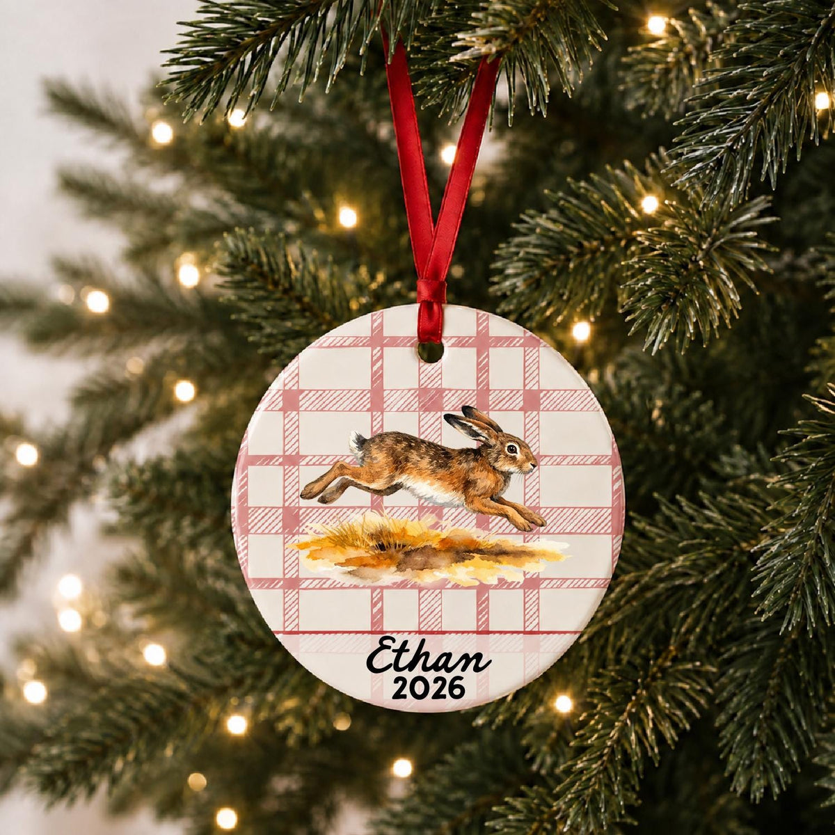 Personalized brown hare ceramic ornament with name and year on red plaid hanging on a lit Christmas tree