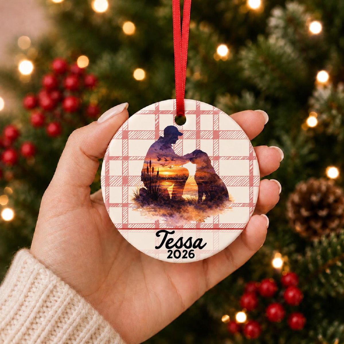 Hand holding the personalized hunter and dog ceramic ornament with name and year on red plaid in front of a Christmas tree