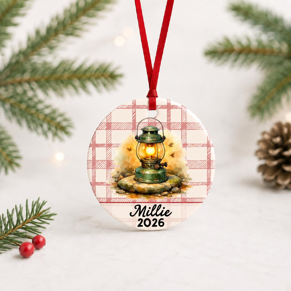 Personalized camping lantern ceramic ornament with name and year on red plaid with red ribbon on a white surface with pine branches and pinecones