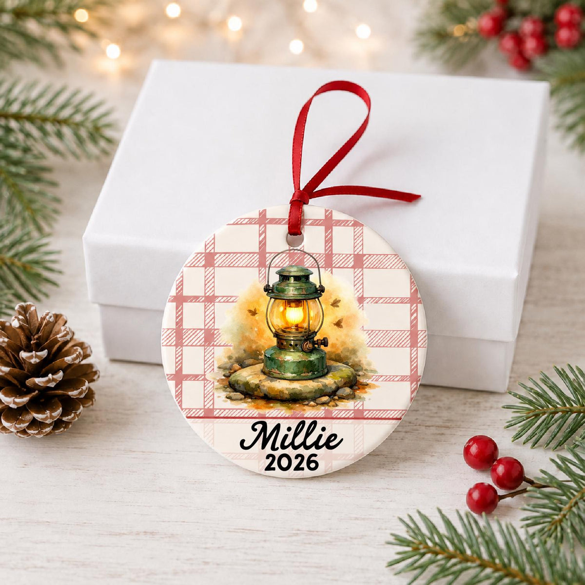 Personalized camping lantern ceramic ornament with name and year on red plaid in front of a white gift box with pinecones and red berries
