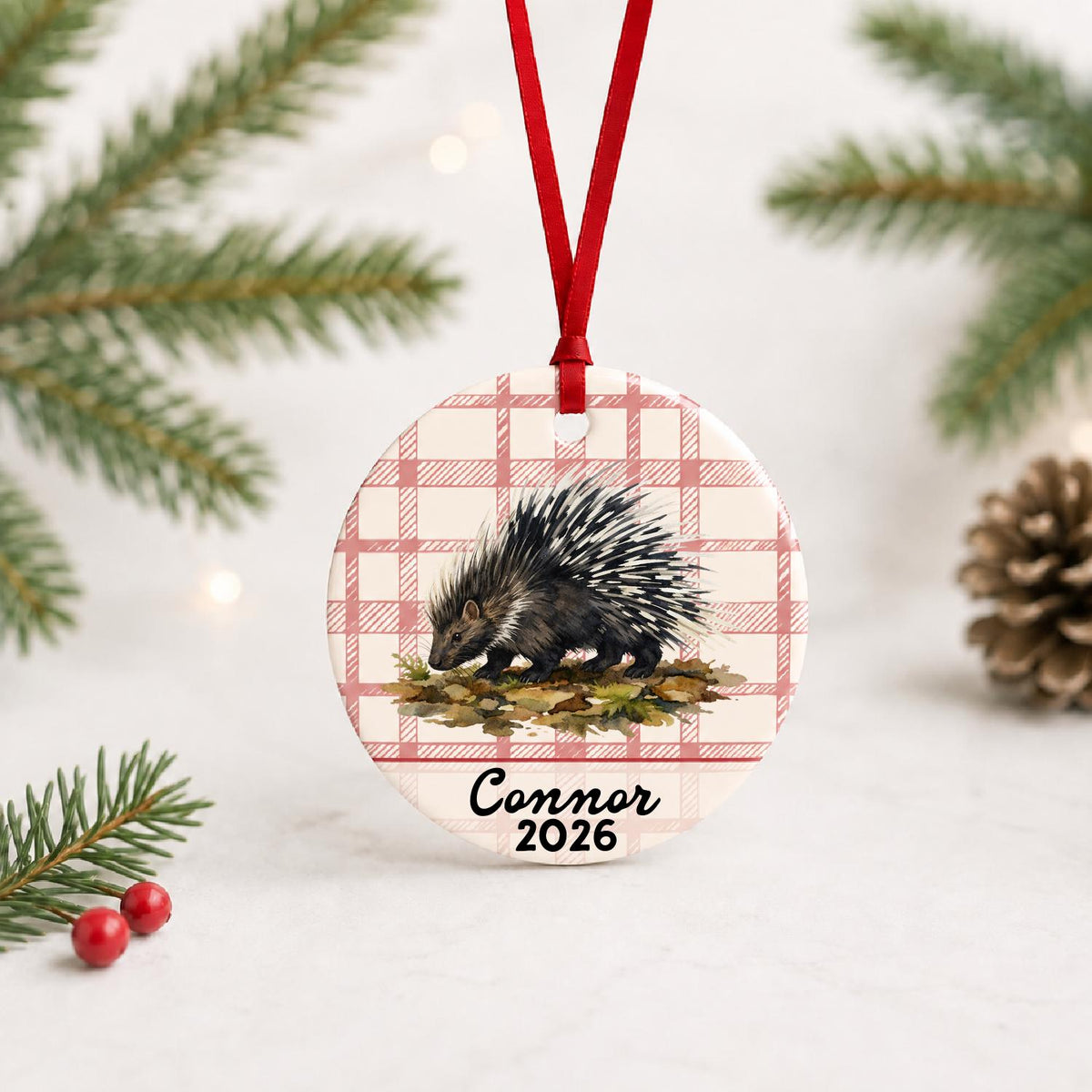 Personalized porcupine ceramic ornament with name and year on red plaid with red ribbon on a white surface with pine branches and pinecones