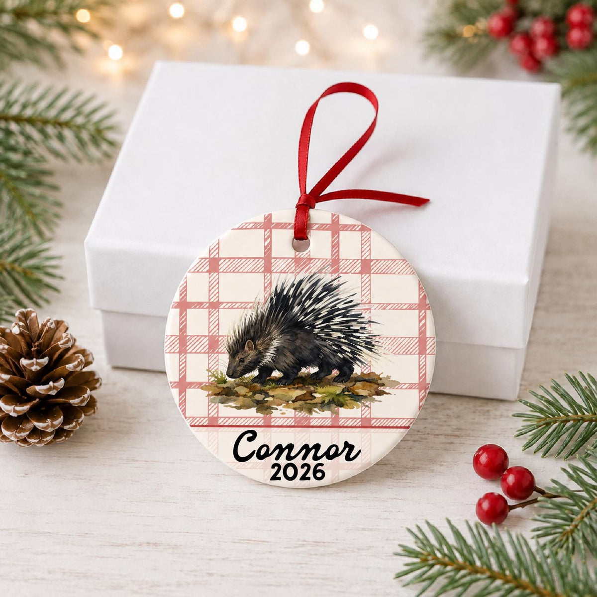 Personalized porcupine ceramic ornament with name and year on red plaid in front of a white gift box with pinecones and red berries