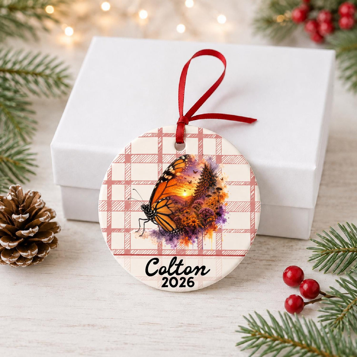 Personalized monarch butterfly ceramic ornament with name and year on red plaid in front of a white gift box with pinecones and red berries