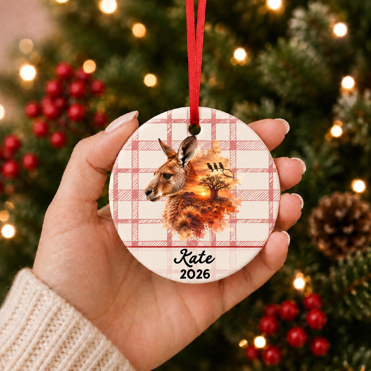Hand holding the personalized red kangaroo ceramic ornament with name and year on red plaid in front of a Christmas tree