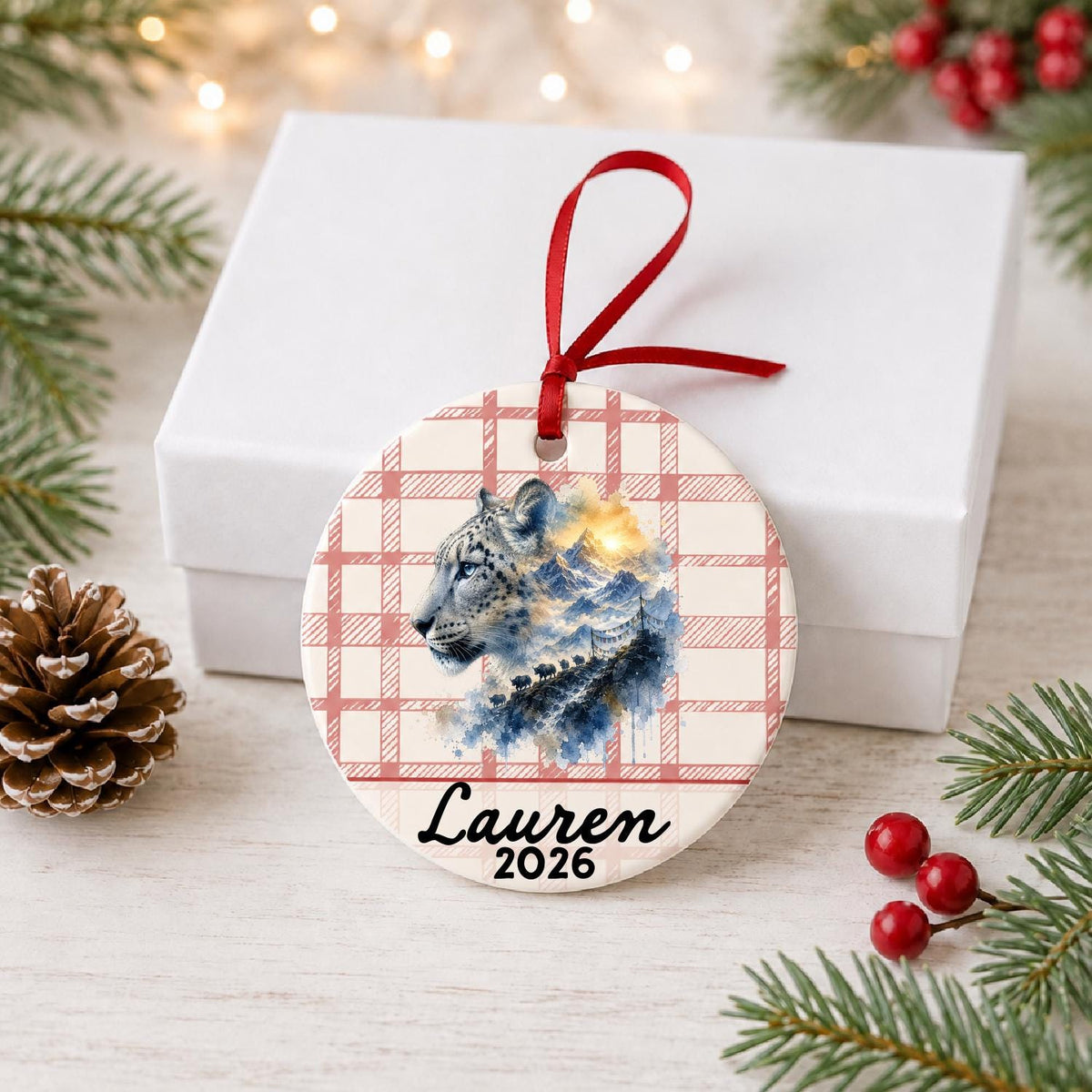 Personalized snow leopard ceramic ornament with name and year on red plaid in front of a white gift box with pinecones and red berries