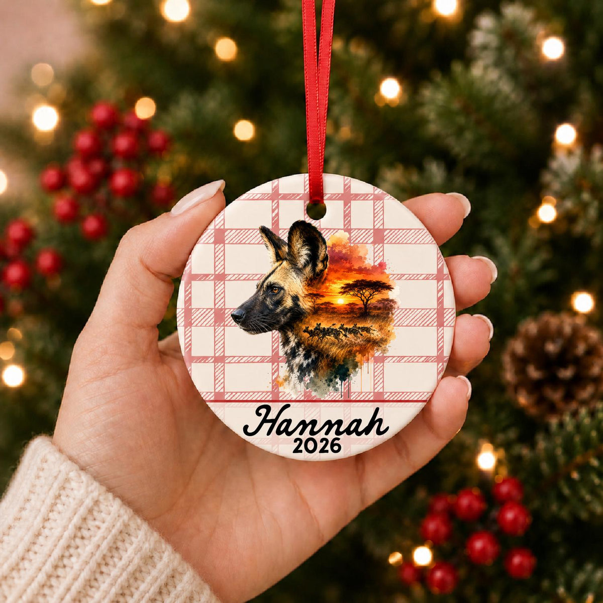 Hand holding the personalized african wild dog ceramic ornament with name and year on red plaid in front of a Christmas tree