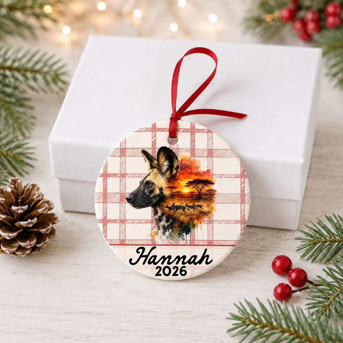 Personalized african wild dog ceramic ornament with name and year on red plaid in front of a white gift box with pinecones and red berries