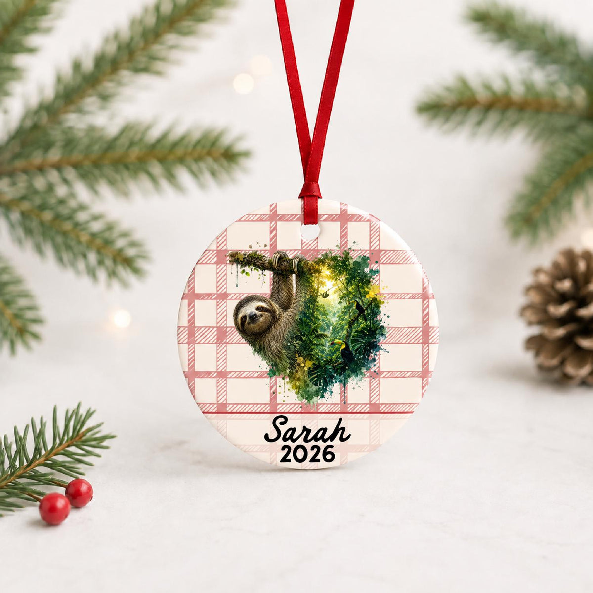 Personalized sloth ceramic ornament with name and year on red plaid with red ribbon on a white surface with pine branches and pinecones