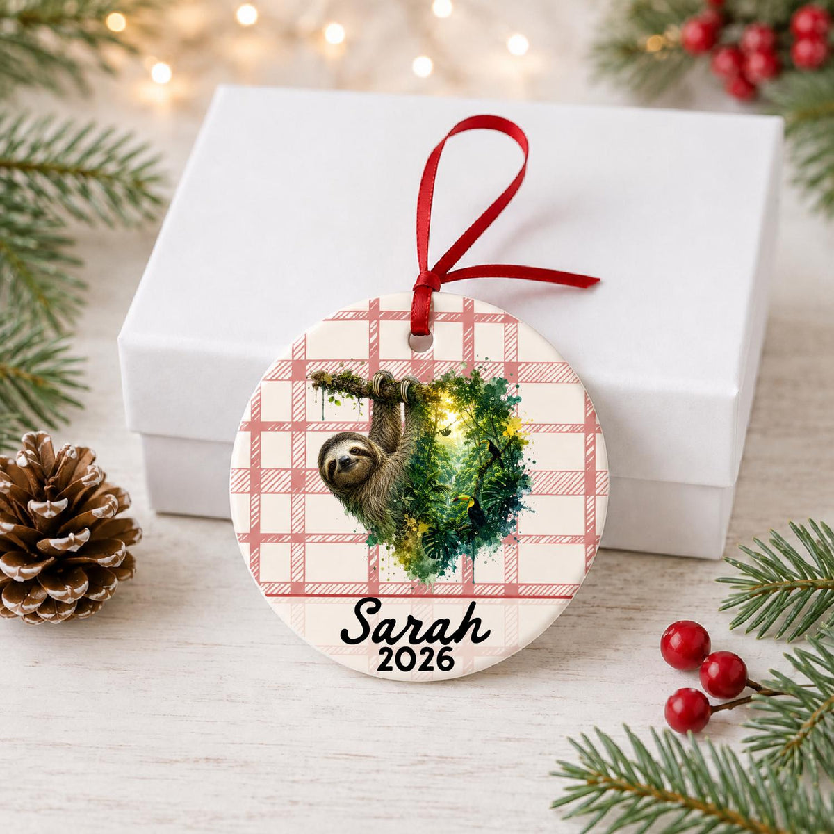 Personalized sloth ceramic ornament with name and year on red plaid in front of a white gift box with pinecones and red berries