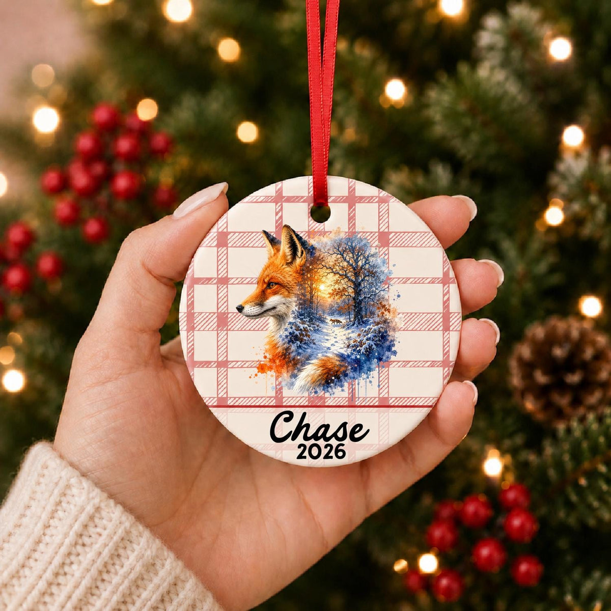 Hand holding the personalized red fox ceramic ornament with name and year on red plaid in front of a Christmas tree