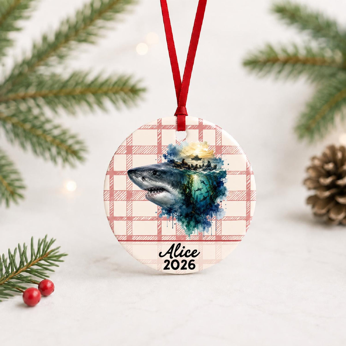 Personalized great white shark ceramic ornament with name and year on red plaid with red ribbon on a white surface with pine branches and pinecones