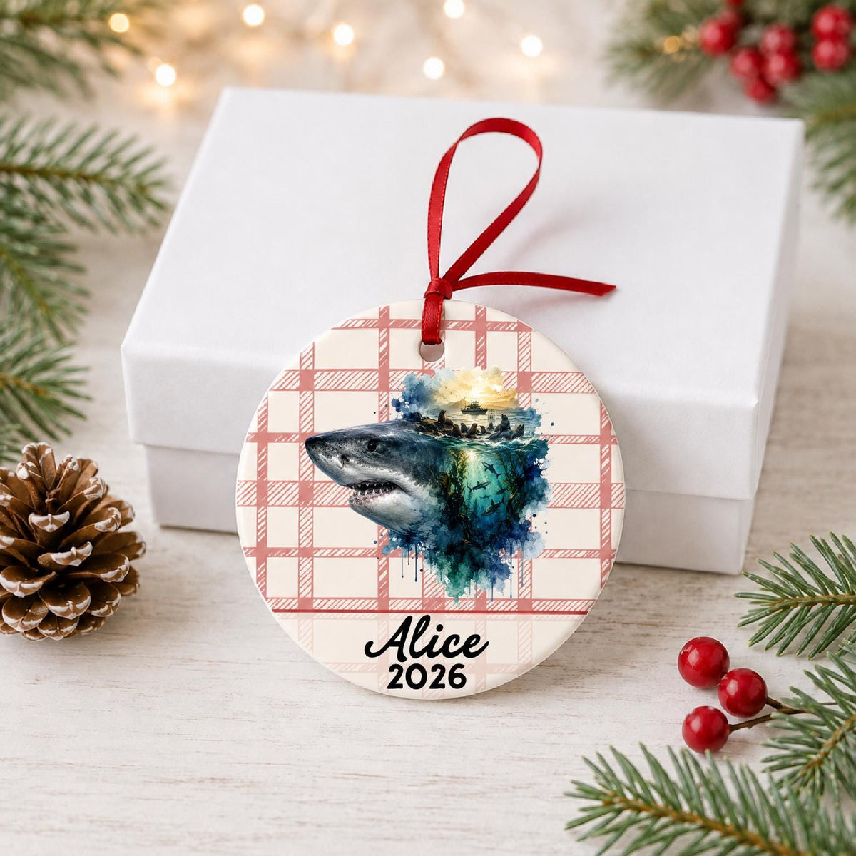 Personalized great white shark ceramic ornament with name and year on red plaid in front of a white gift box with pinecones and red berries