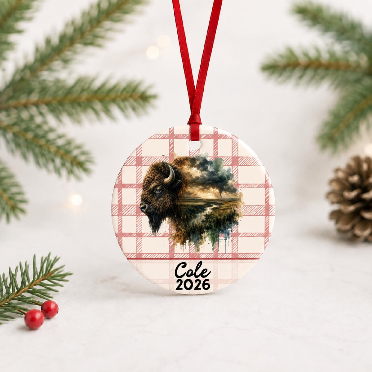 Personalized american bison ceramic ornament with name and year on red plaid with red ribbon on a white surface with pine branches and pinecones