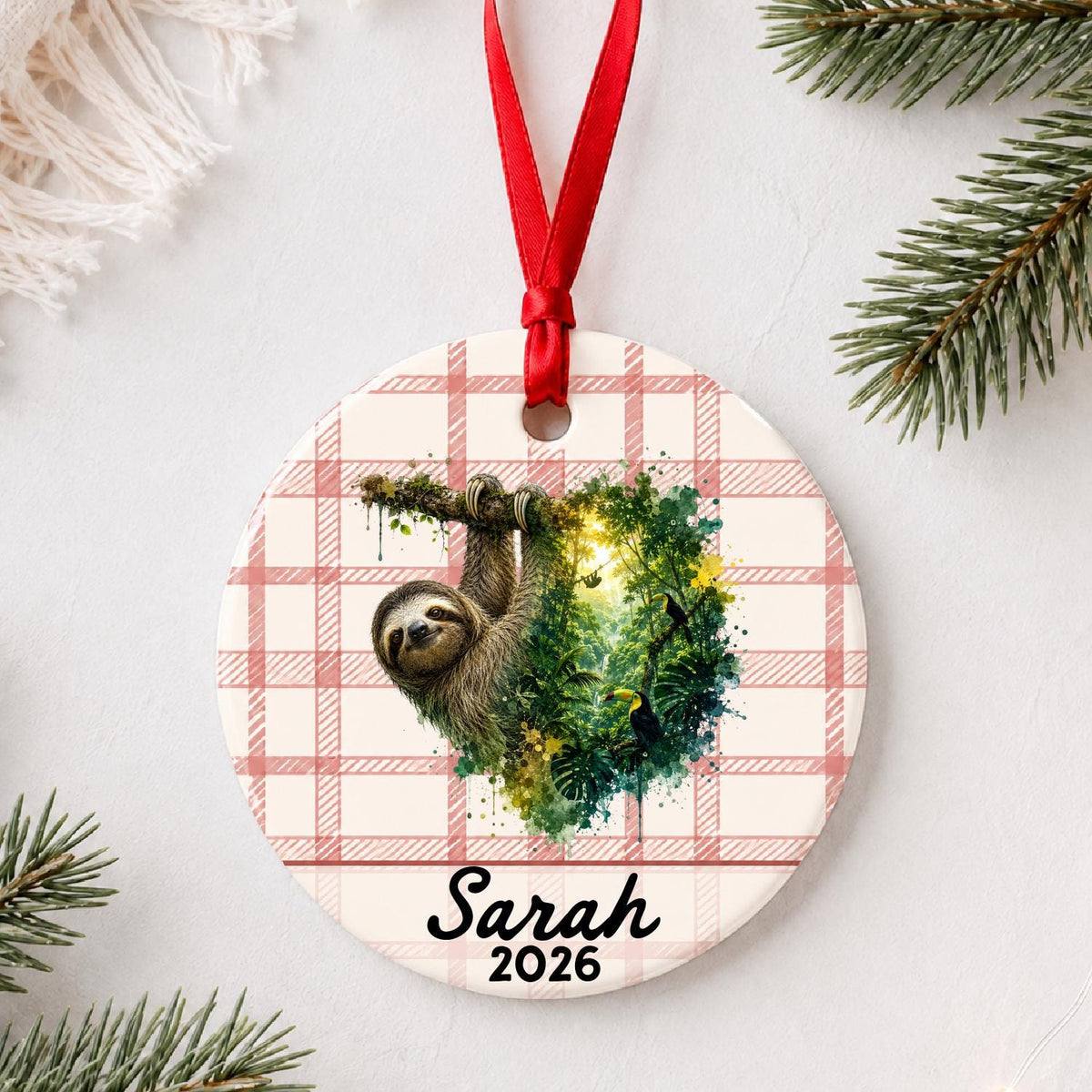 Personalized sloth ceramic ornament with name and year on red plaid styled with pine branches on a soft white background