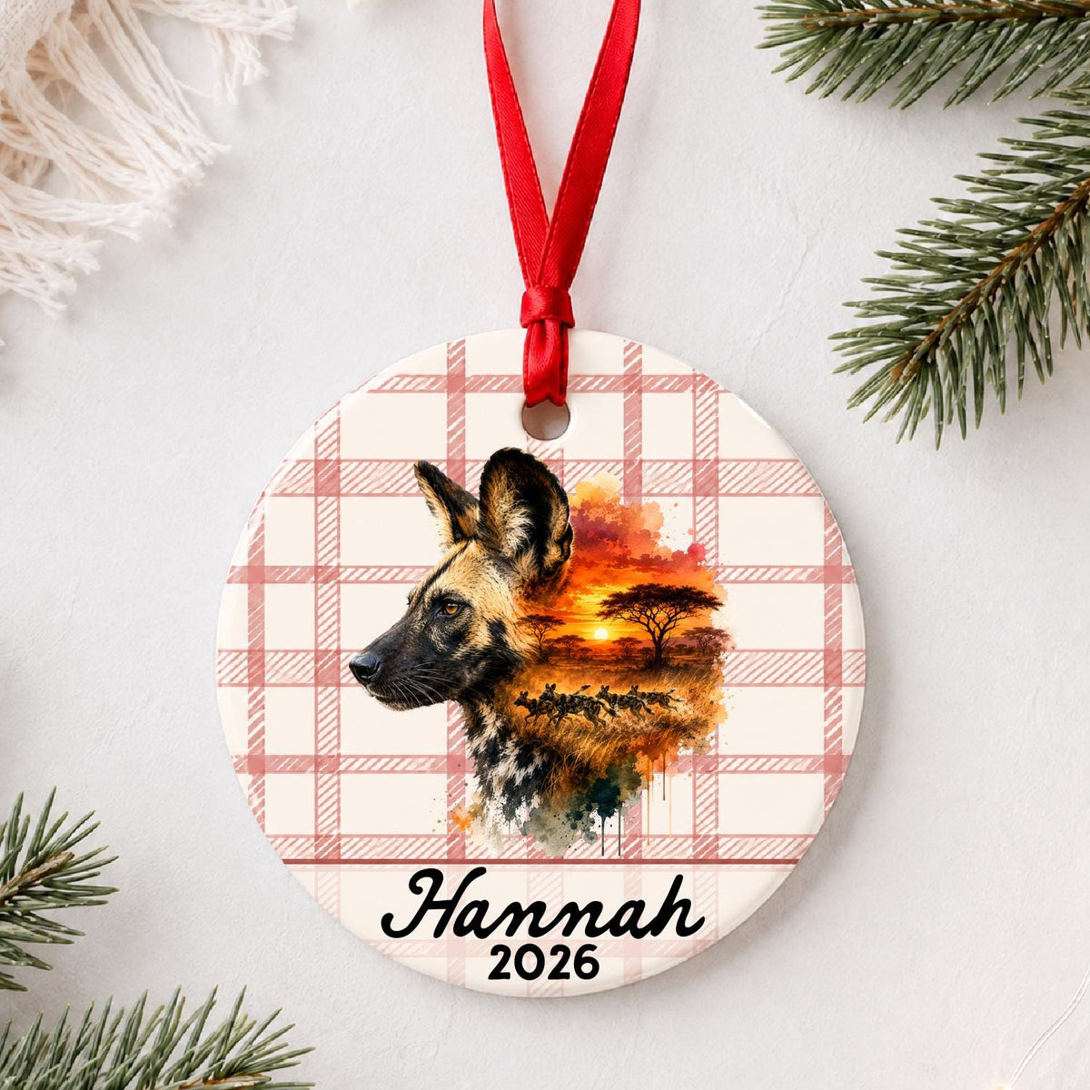 Personalized african wild dog ceramic ornament with name and year on red plaid styled with pine branches on a soft white background