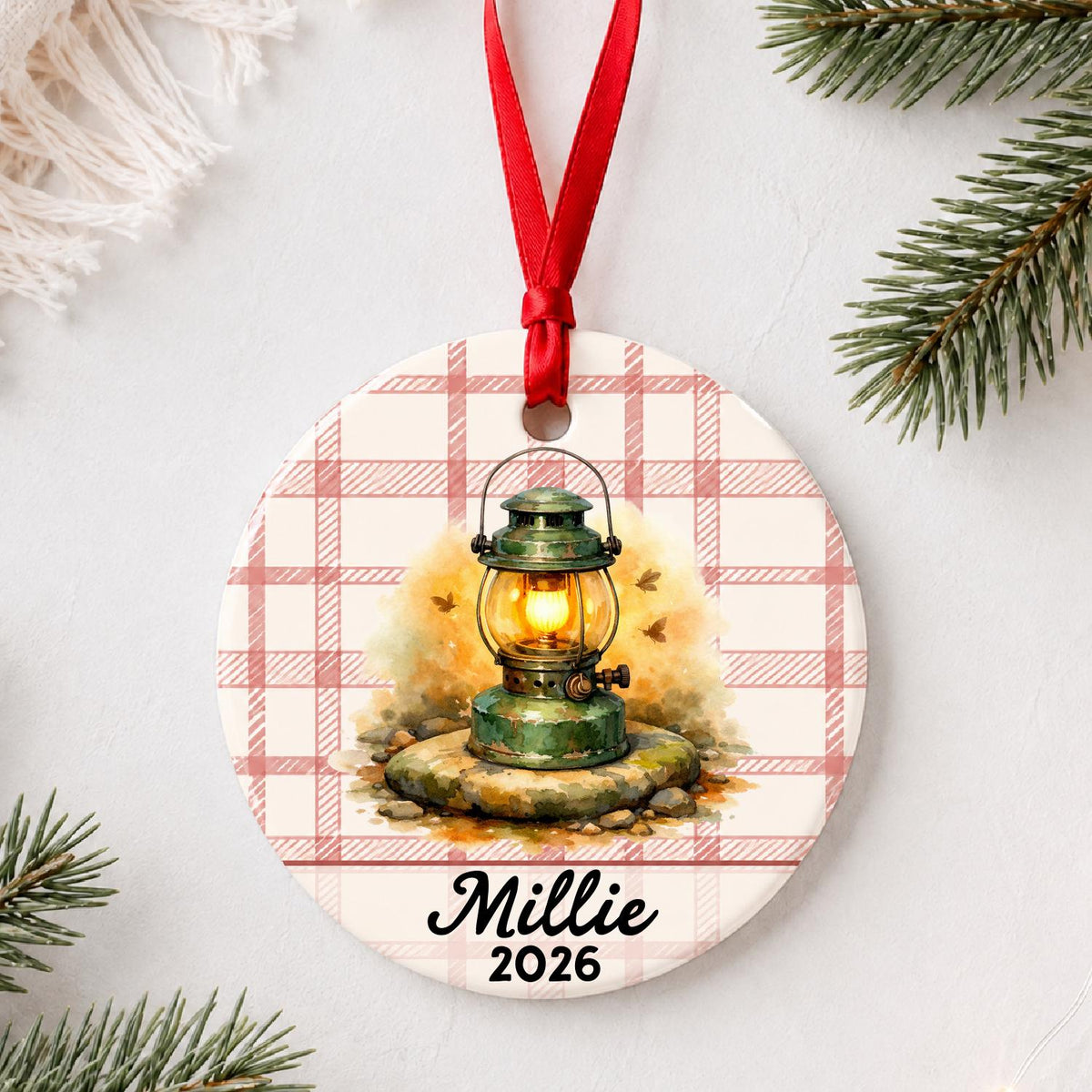 Personalized camping lantern ceramic ornament with name and year on red plaid styled with pine branches on a soft white background