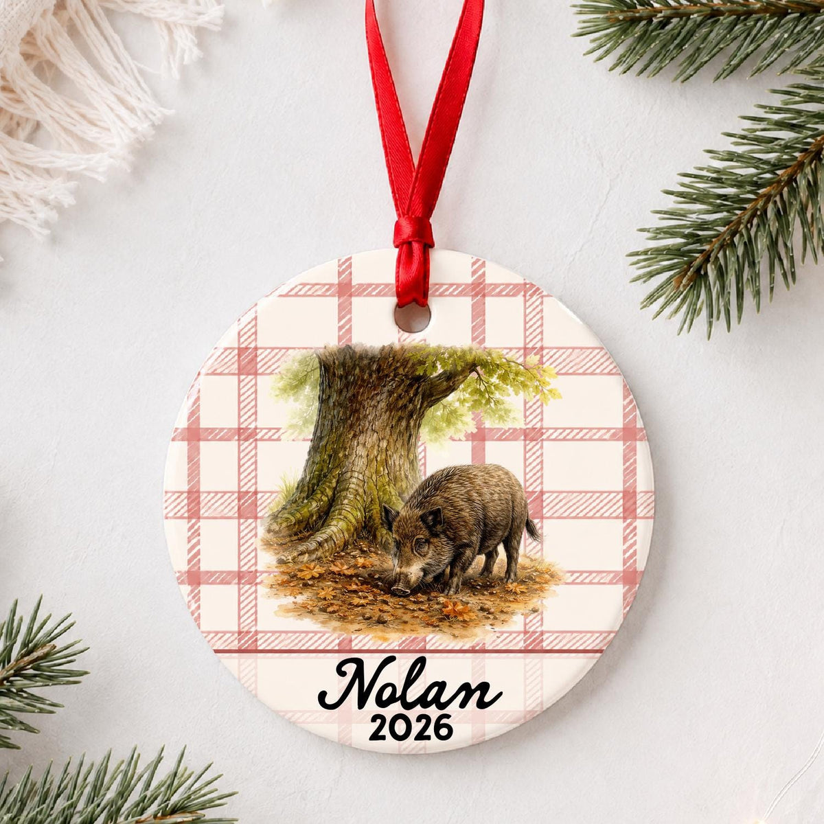 Personalized wild boar ceramic ornament with name and year on red plaid styled with pine branches on a soft white background