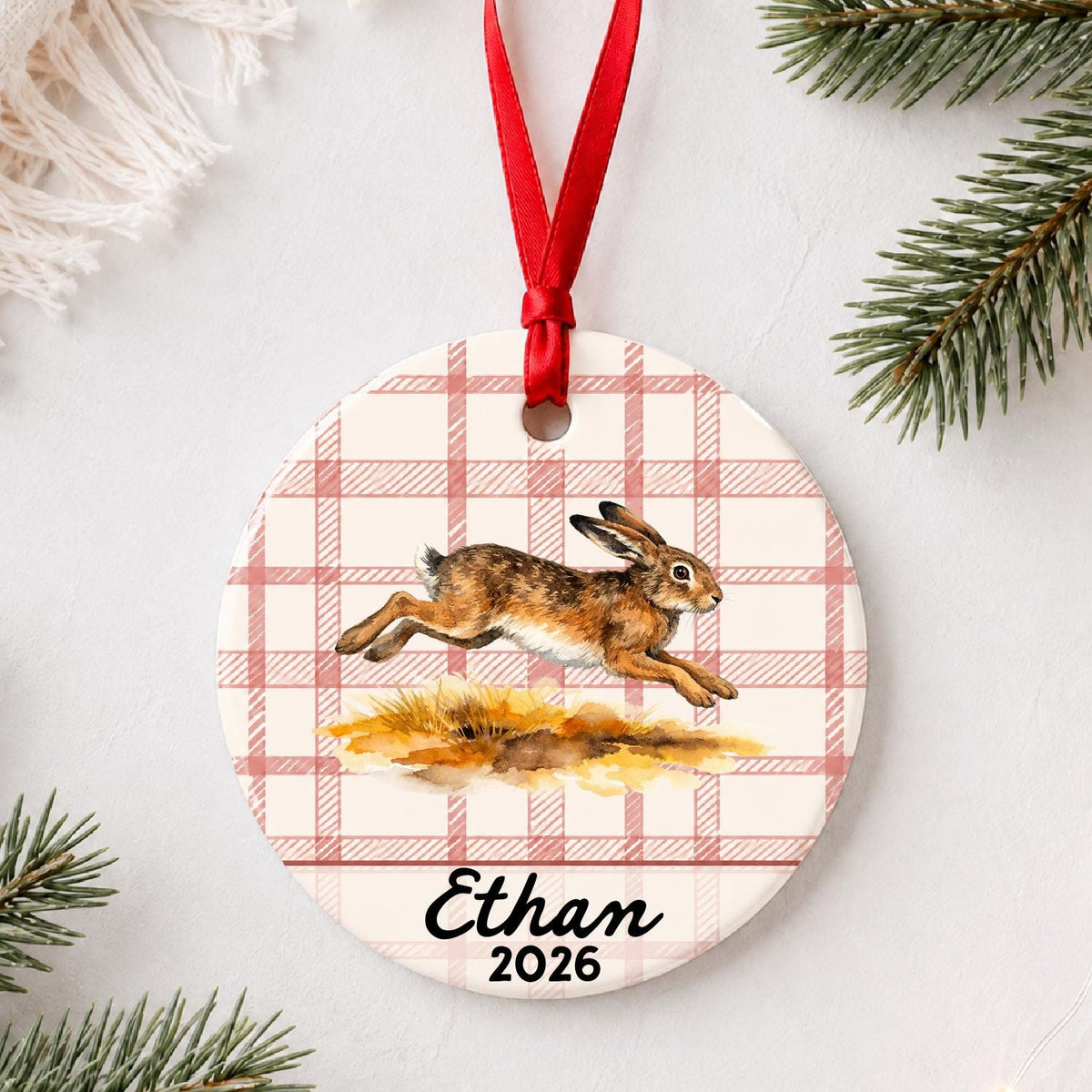 Personalized brown hare ceramic ornament with name and year on red plaid styled with pine branches on a soft white background